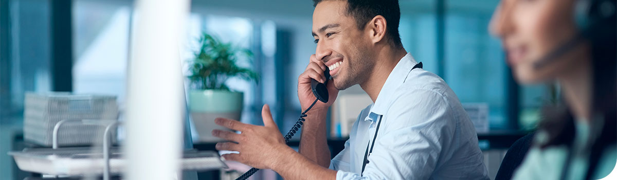 Un homme souriant discute au téléphone à son bureau de travail en regardant son écran d'ordinateur